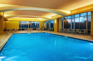 An indoor swimming pool with clear blue water, surrounded by chairs and tables, and large windows offering a view of the outdoors.