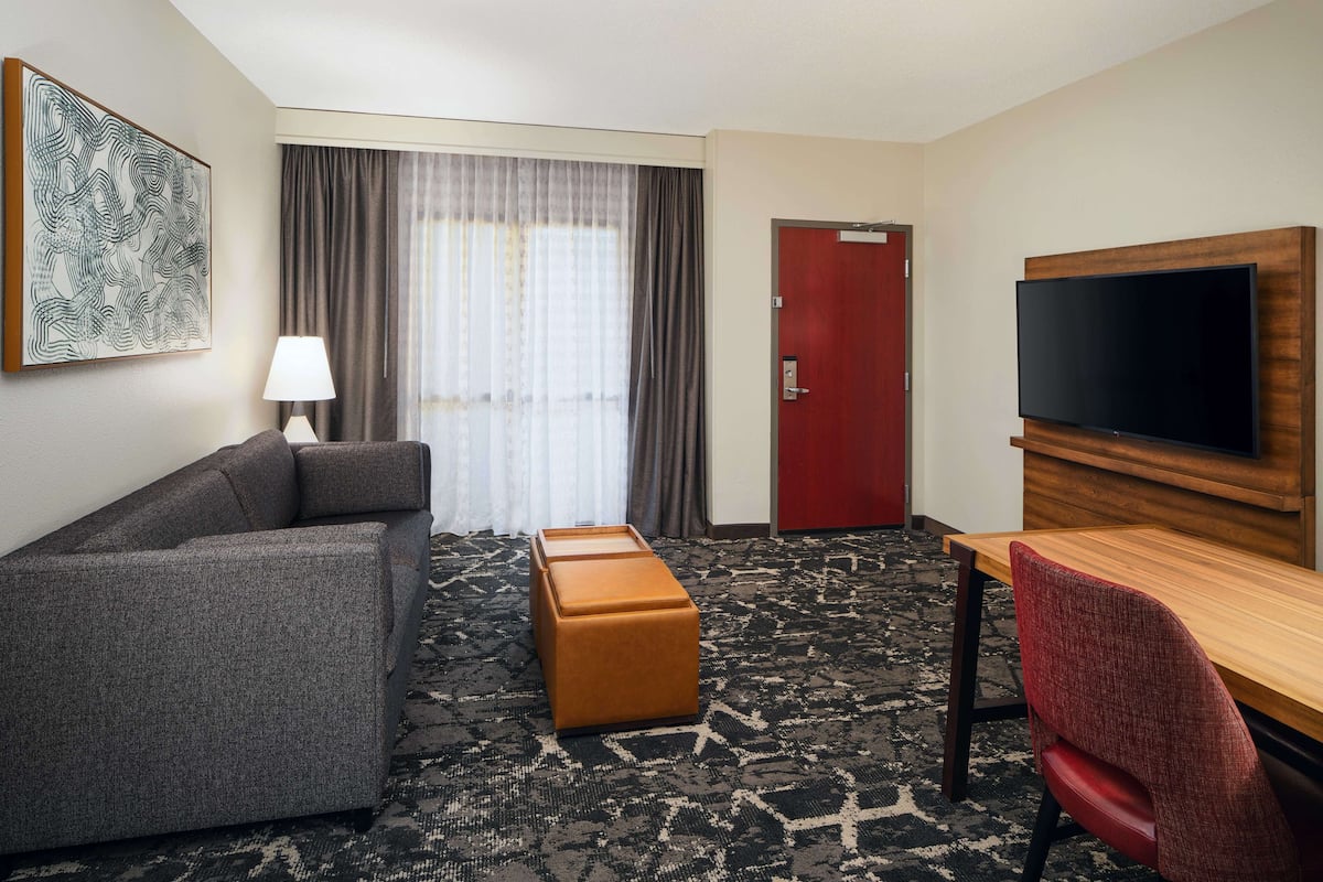 A hotel room with a sofa, a wooden desk, a television, a red door, and a window with curtains.