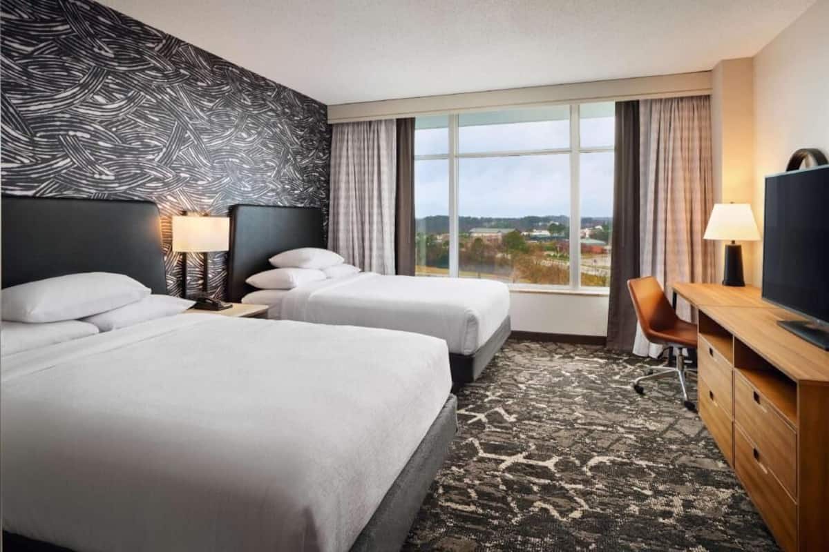 A hotel room with two beds, a desk, a TV, and a large window with a view.