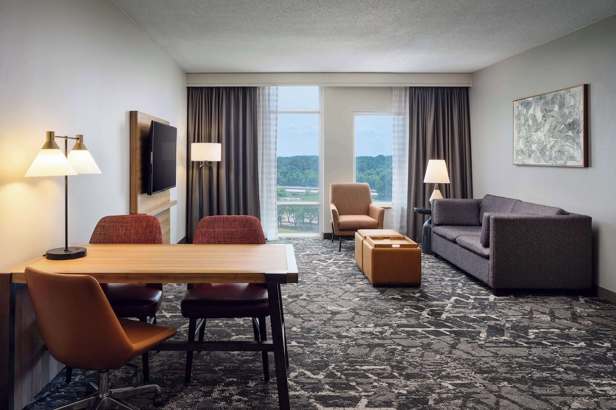 A hotel room with a large window, a flat-screen TV, a sofa, a chair, a table, and lamps.