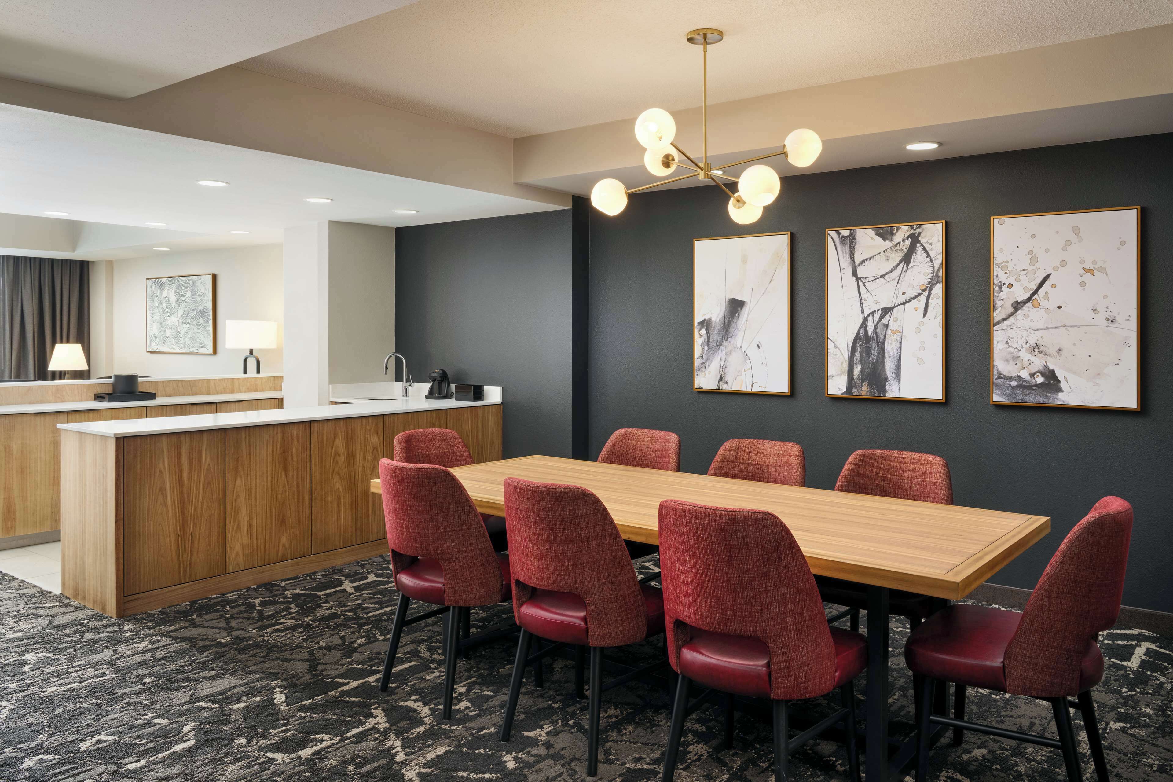 A modern hotel room with a wooden dining table, red upholstered chairs, a black and white abstract painting on the wall, and a black and white abstract painting on the wall.