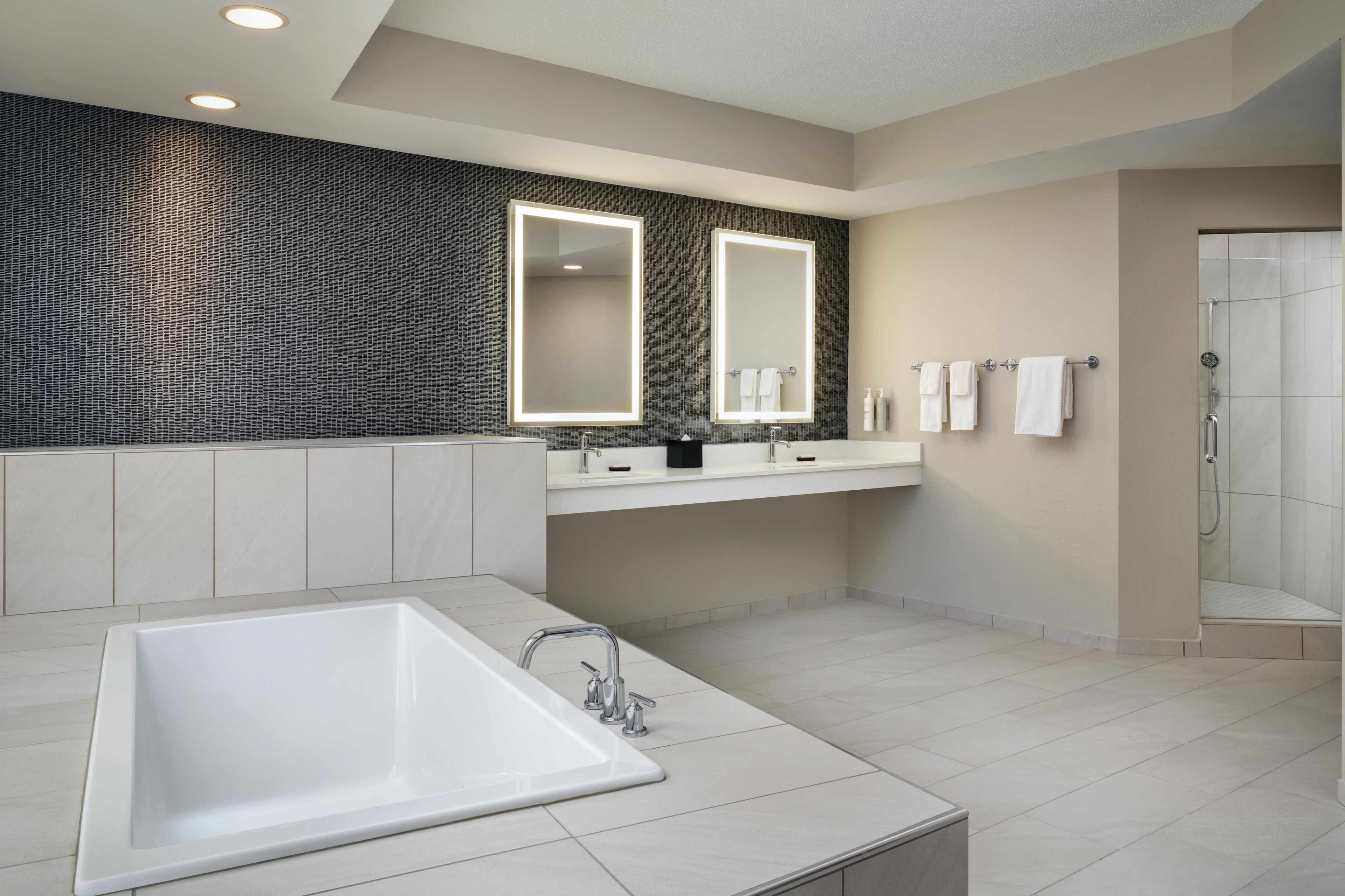 A modern bathroom with a large bathtub, two sinks, a walk-in shower, and a textured wall.