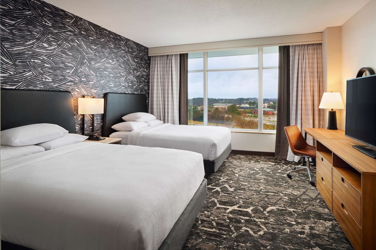 A hotel room with two beds, a desk, a TV, and a large window with a view of the city.