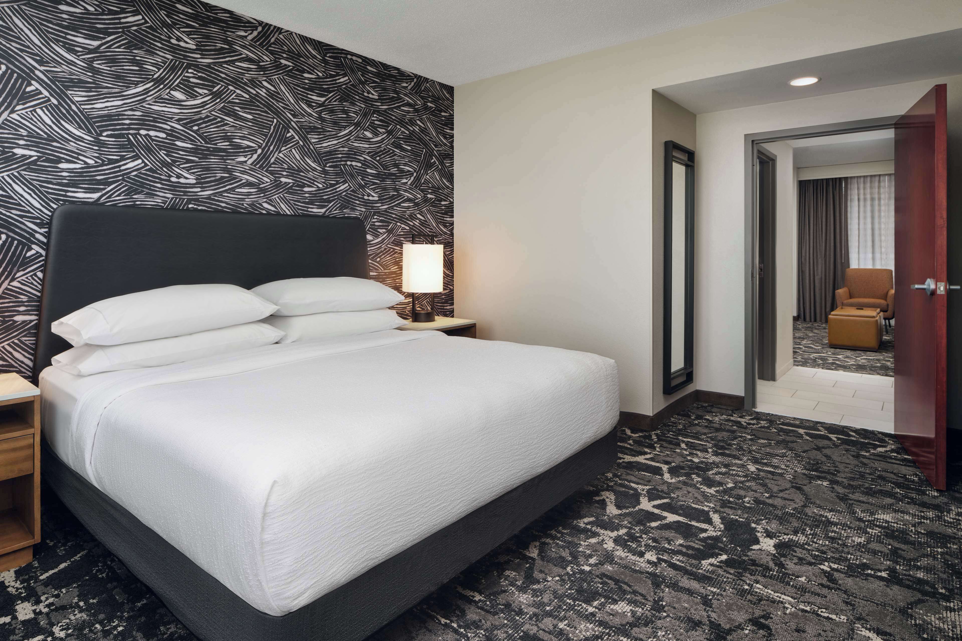 Une chambre d’hôtel avec un grand lit, une table de chevet, une lampe, une moquette à motifs et une porte donnant sur une autre pièce.