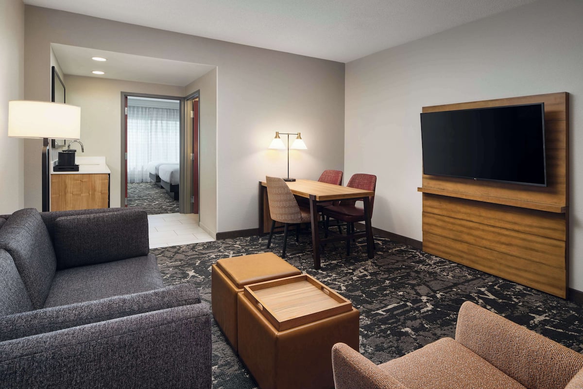 A hotel room with a sofa, a small wooden table, a TV mounted on the wall, a desk with a lamp, and a bed in the background.
