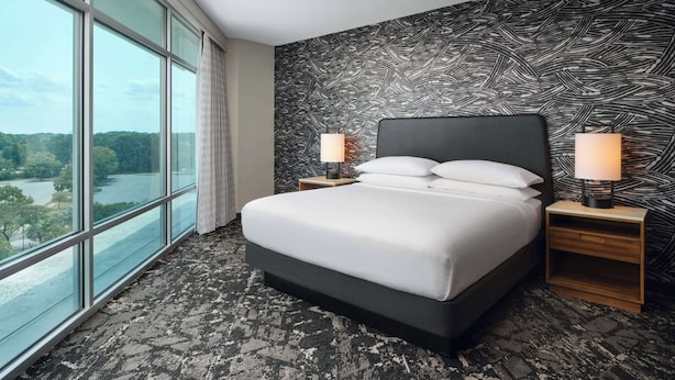 A hotel room with a large window, a bed with white linens, two bedside lamps, and a patterned wall. - Embassy Suites by Hilton Hampton Convention Center (Hampton)