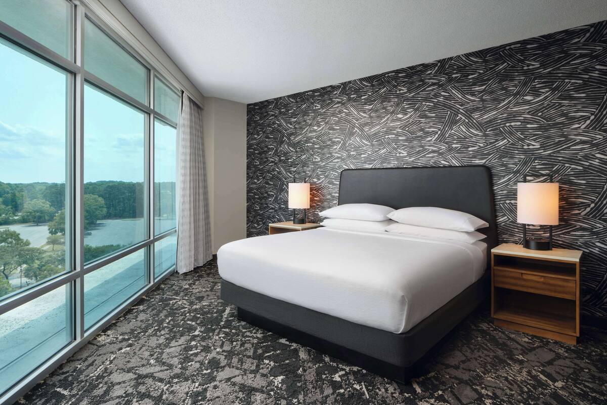 A hotel room with a large window, a bed with white linens, two bedside lamps, and a patterned wall.