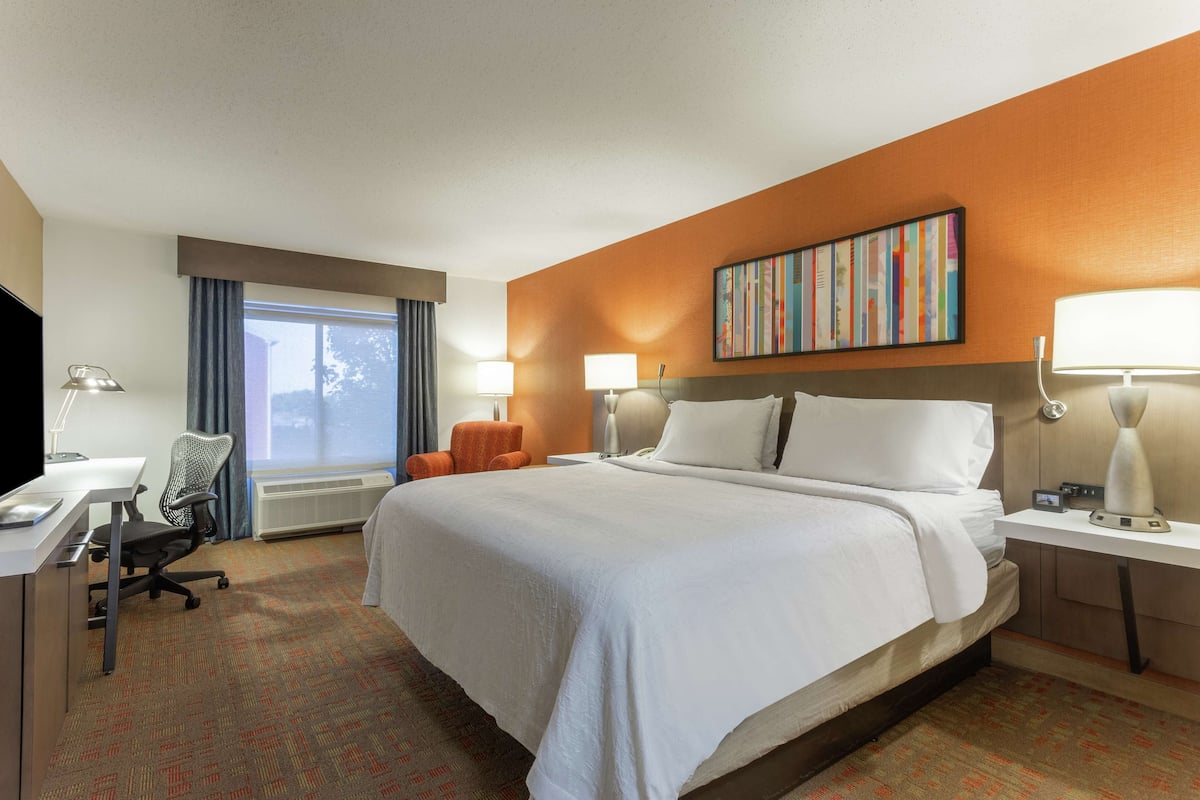 A hotel room with a large bed, two bedside tables with lamps, a desk with a chair, and a large window with curtains.