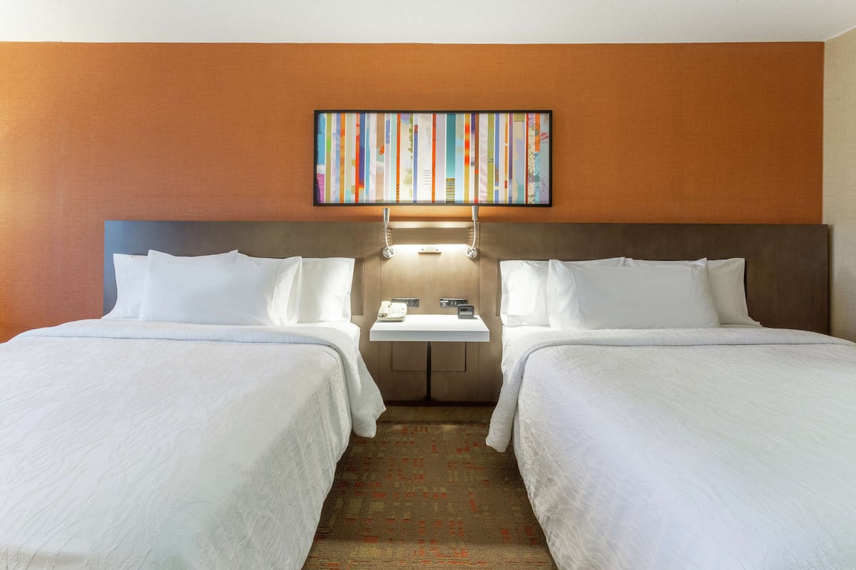 A hotel room with two beds, a small table with a telephone and a clock, and a wall-mounted artwork above the beds.