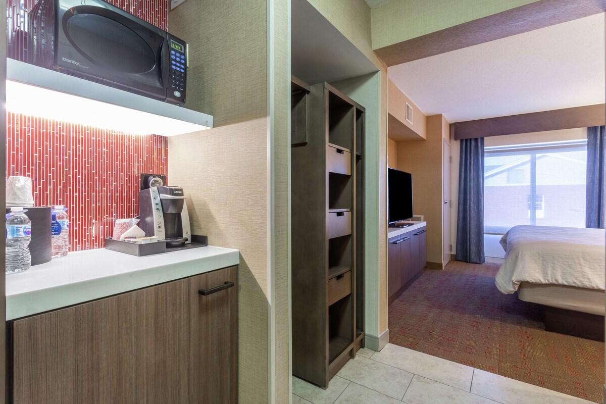 Habitación de hotel con kitchenette, cama, televisor y ventana con cortinas.