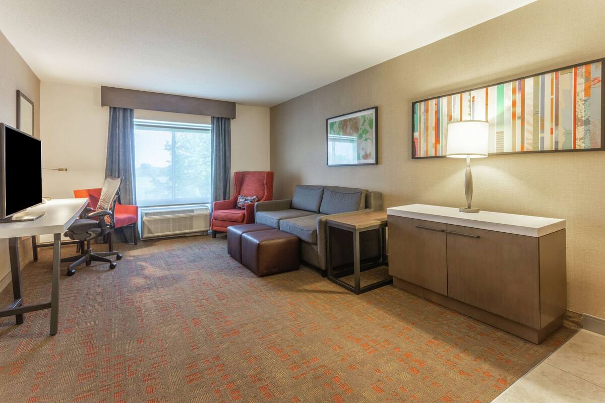 Hotelkamer met bureau, rode fauteuil, grijze bank, klein tafeltje, kast en groot raam met gordijnen.