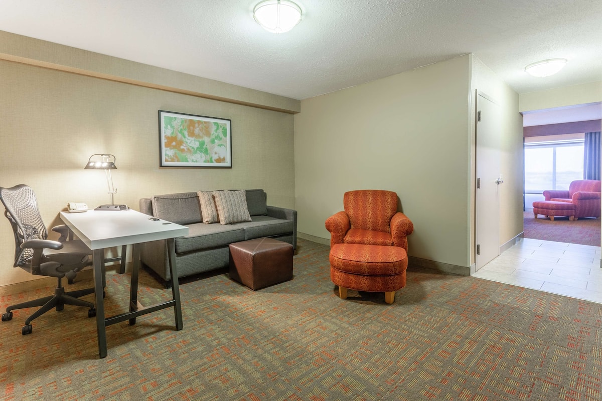 Een hotelkamer met een bureau, bureaustoel, bank, poef en een oranje fauteuil.