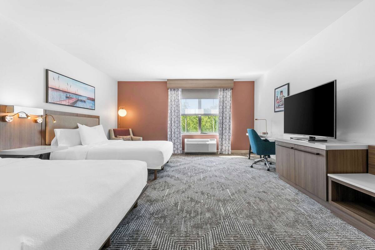 A hotel room with two beds, a desk, a TV, and a large window with curtains.