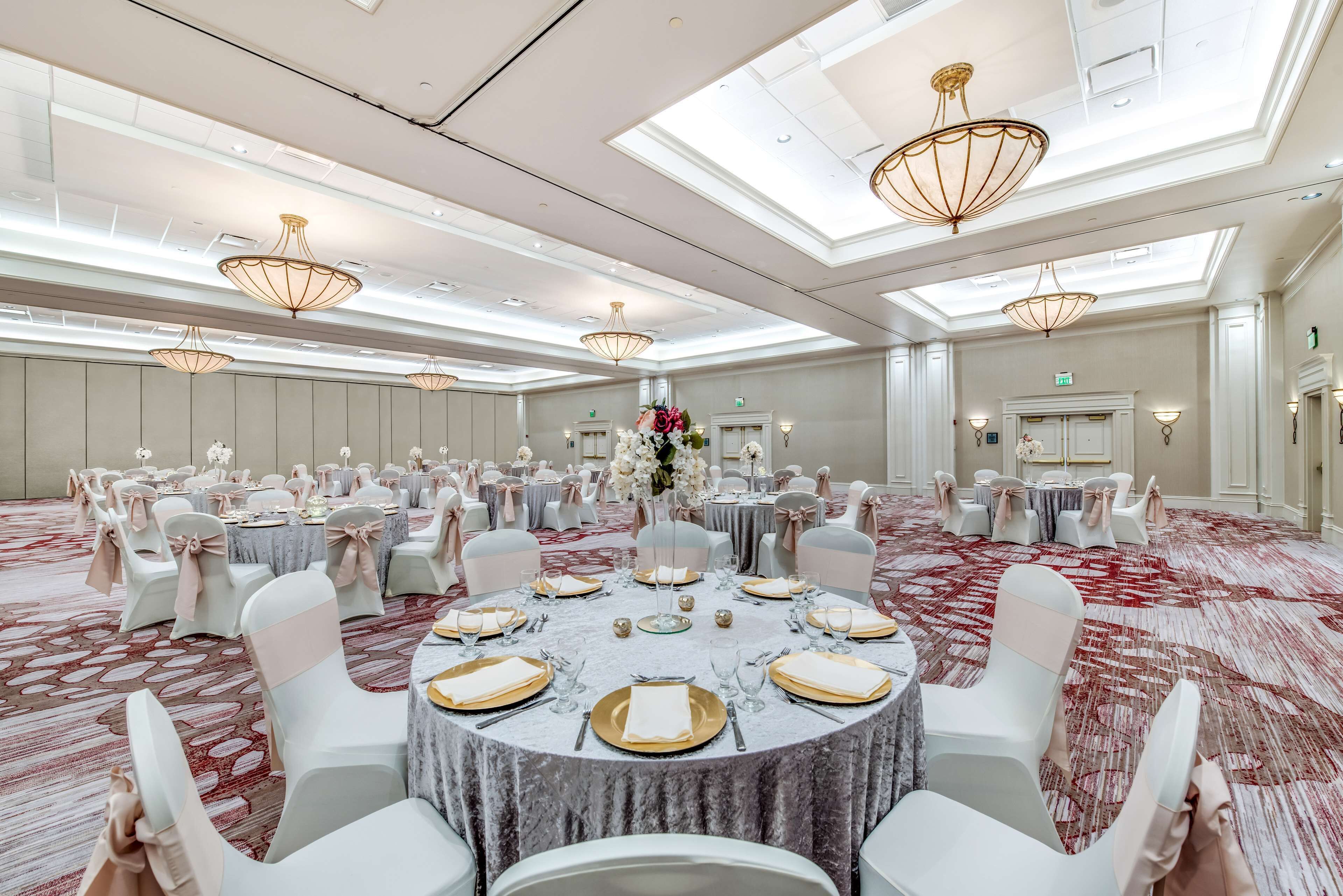 Uma sala de banquetes com mesas redondas preparadas para refeições, cadeiras brancas e lustres.