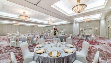 Uma sala de banquetes com mesas redondas preparadas para refeições, cadeiras brancas e lustres.