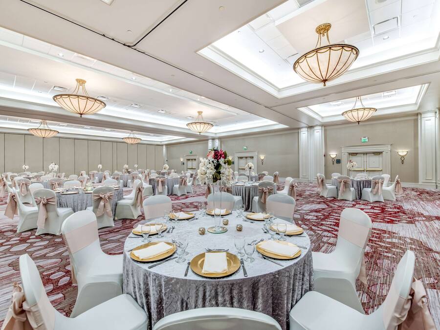 Uma sala de banquetes com mesas redondas preparadas para refeições, cadeiras brancas e lustres.