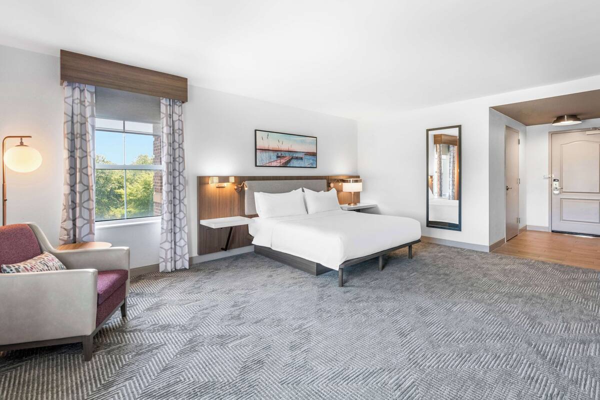 A hotel room with a large bed, a chair, a desk, and a window with curtains.