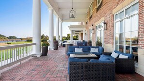 A covered outdoor seating area with brick flooring, white columns, and blue furniture. - Hilton Garden Inn Suffolk Riverfront (Suffolk)