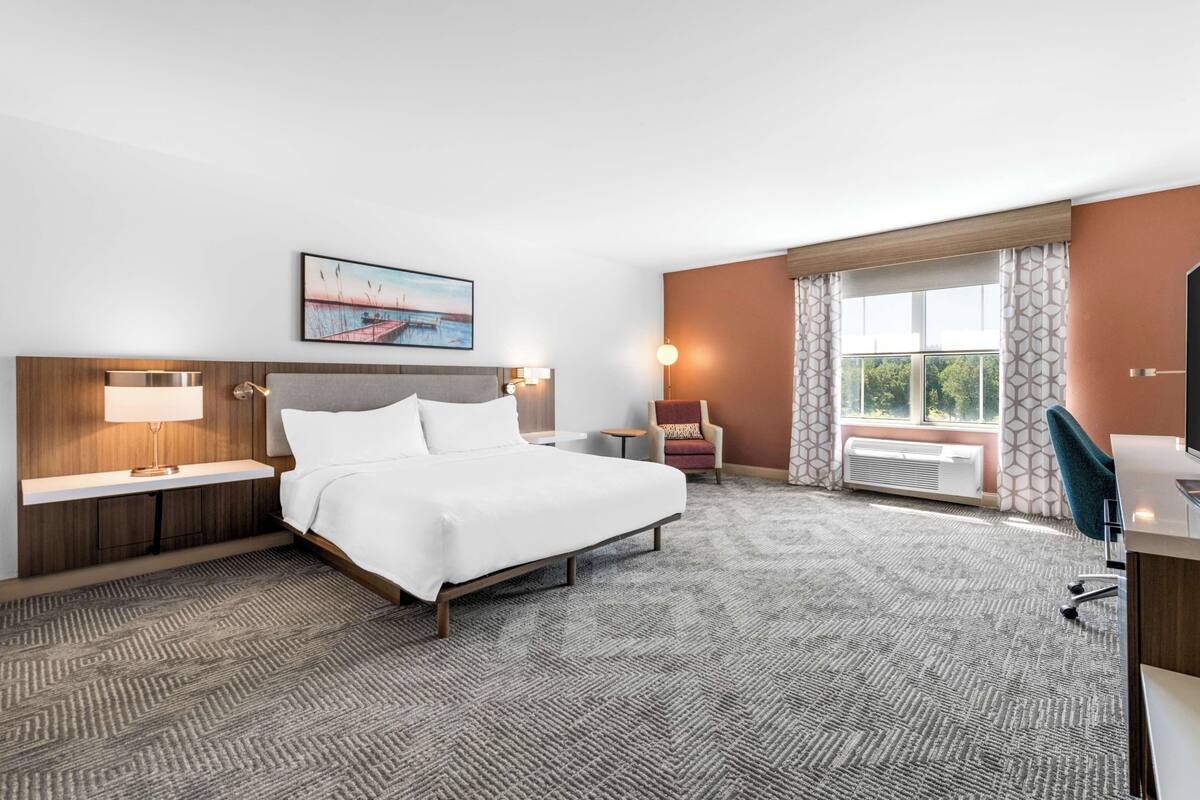 A hotel room with a large bed, a desk, a chair, a small table, and a window with curtains.