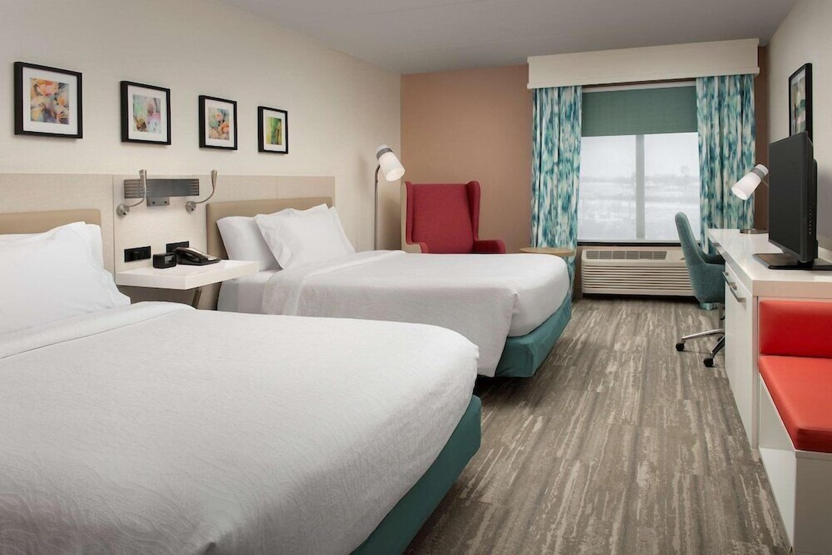 A hotel room with two beds, a desk with a computer, a chair, a TV, and a window with curtains.