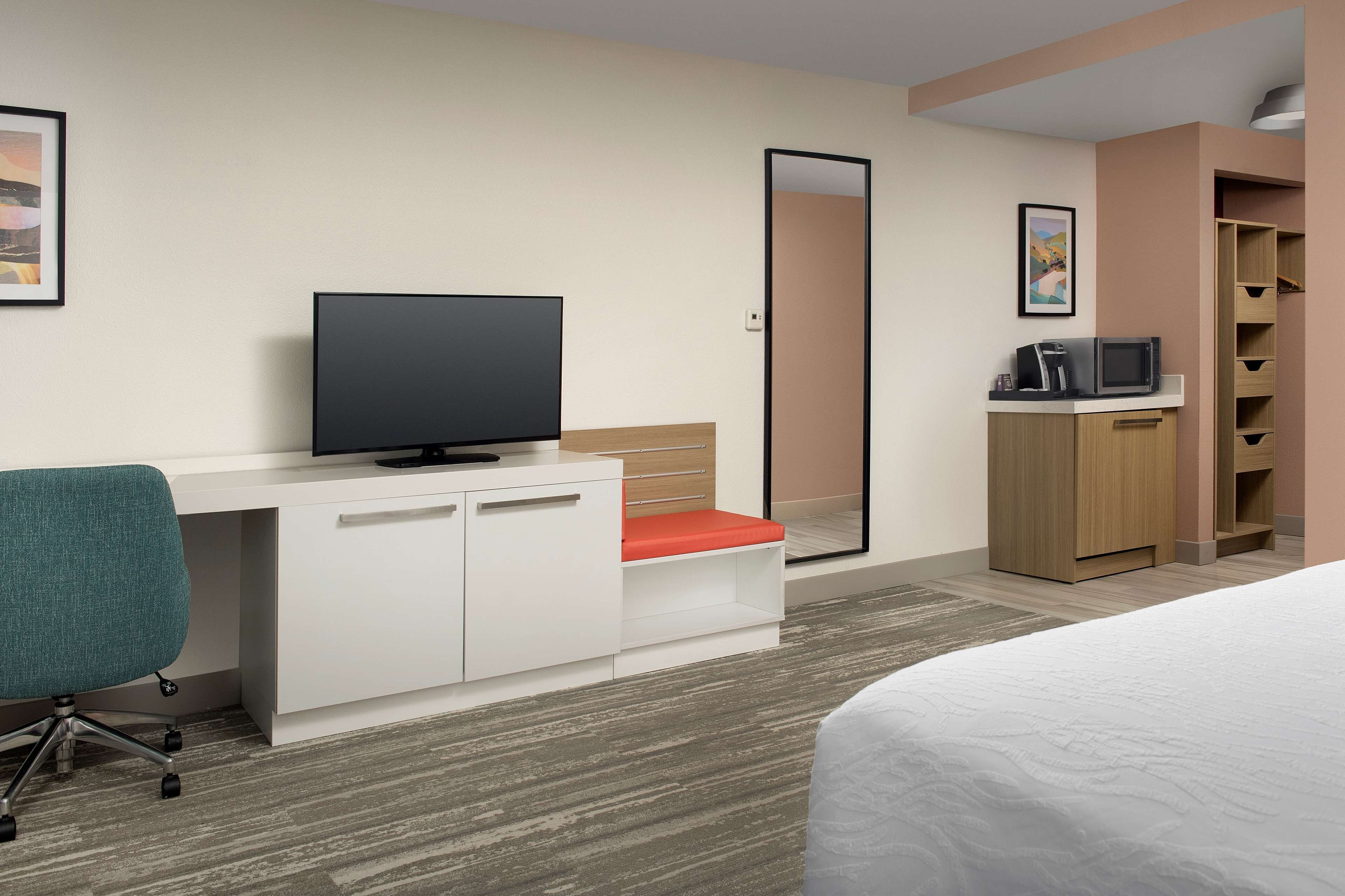 A hotel room with a bed, a desk with a TV, a chair, a wardrobe, and a mirror.