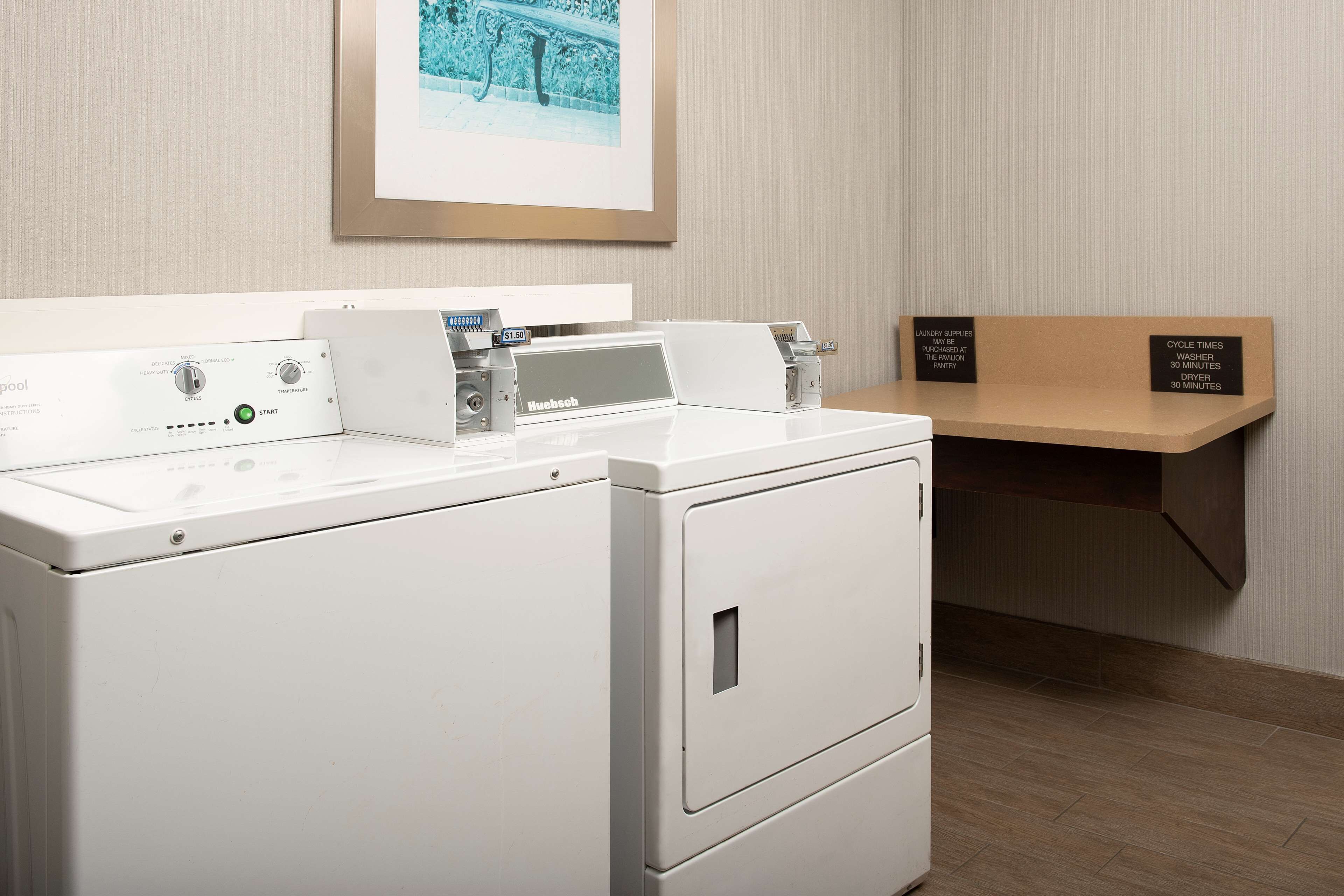 A modern laundry area with a front-loading washer and dryer, a small shelf with a sign indicating cycle times, and a framed picture on the wall.