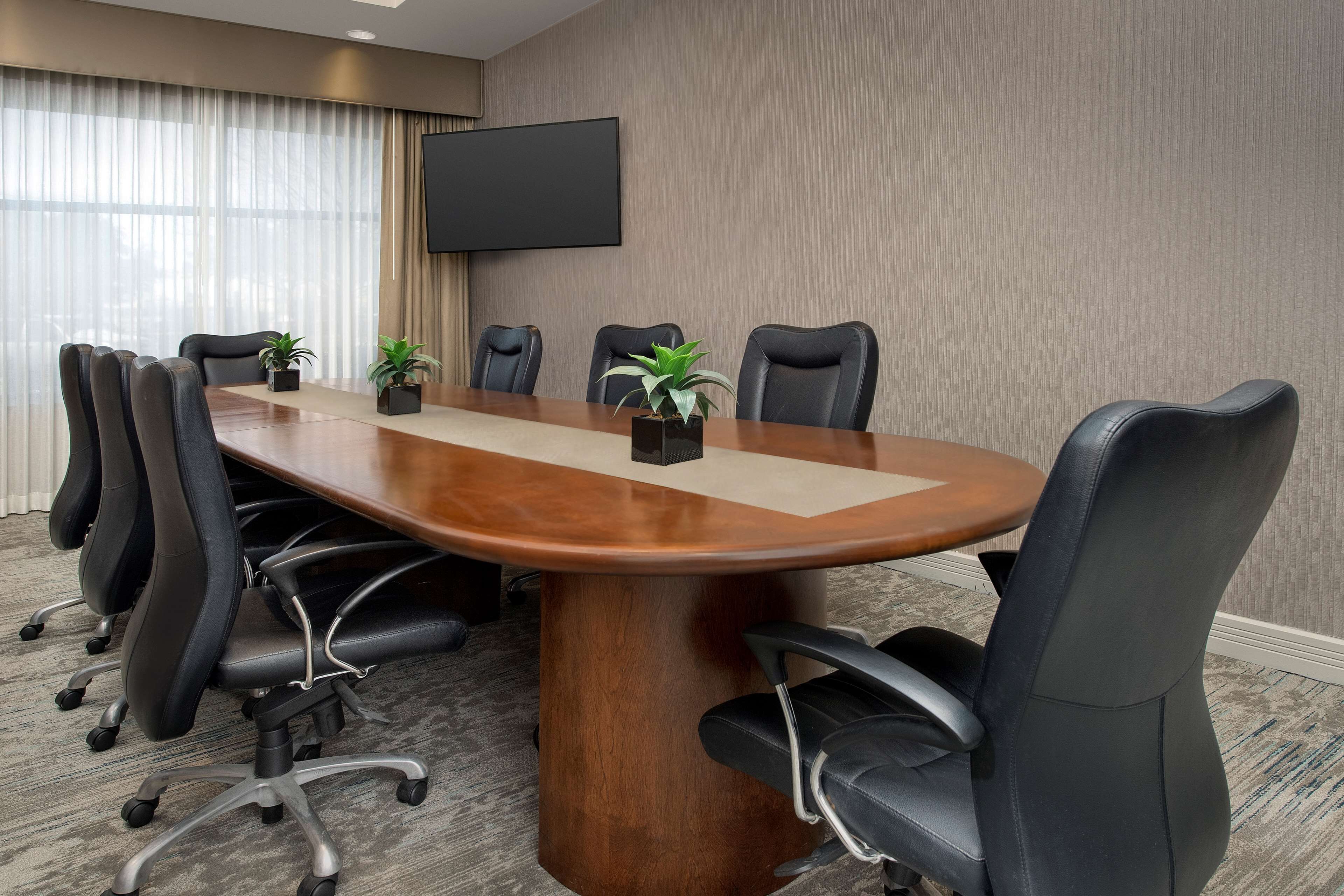 Une salle de conférence avec une grande table en bois, des fauteuils en cuir noir et un téléviseur à écran plat fixé au mur.
