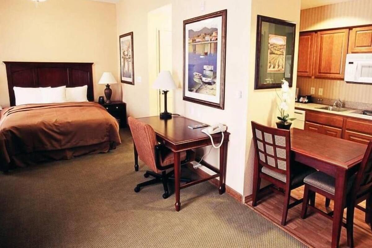 A hotel room with a bed, desk, chair, lamp, and a kitchenette.