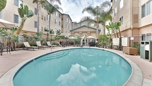 A swimming pool surrounded by lounge chairs and palm trees, with a multi-story building in the background.