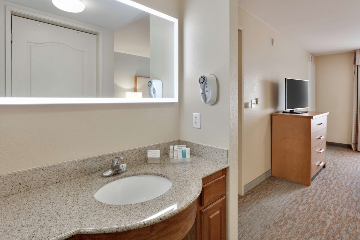 A hotel bathroom with a granite countertop sink, a mirror, a television on a wooden cabinet, and a door leading to another room.