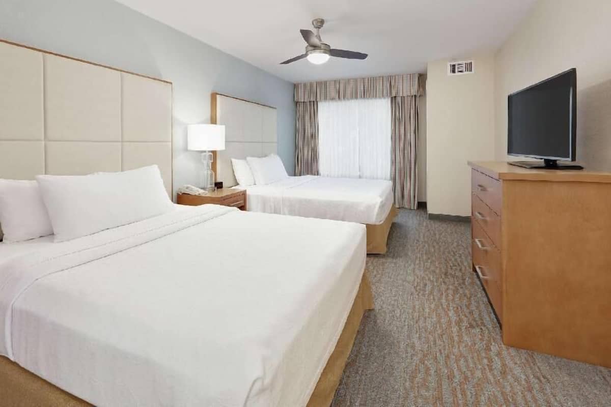 A hotel room with two beds, a TV, a desk, a lamp, and a ceiling fan.