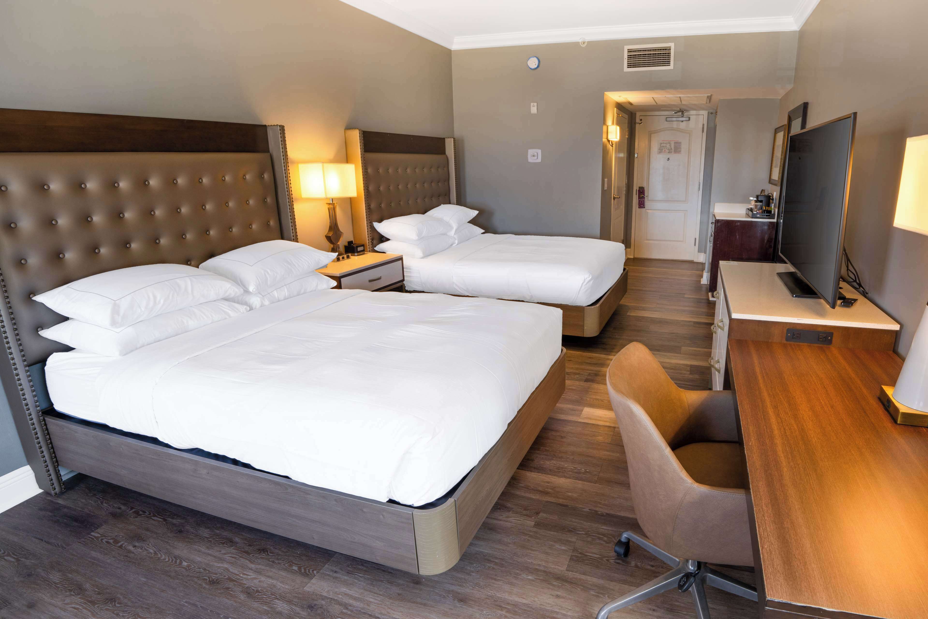 A hotel room with two beds, a desk, a chair, a TV, and a lamp.