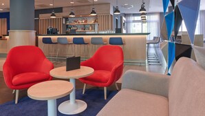 Un moderno vestíbulo de hotel con asientos rojos y grises, una zona de bar y una escalera.
