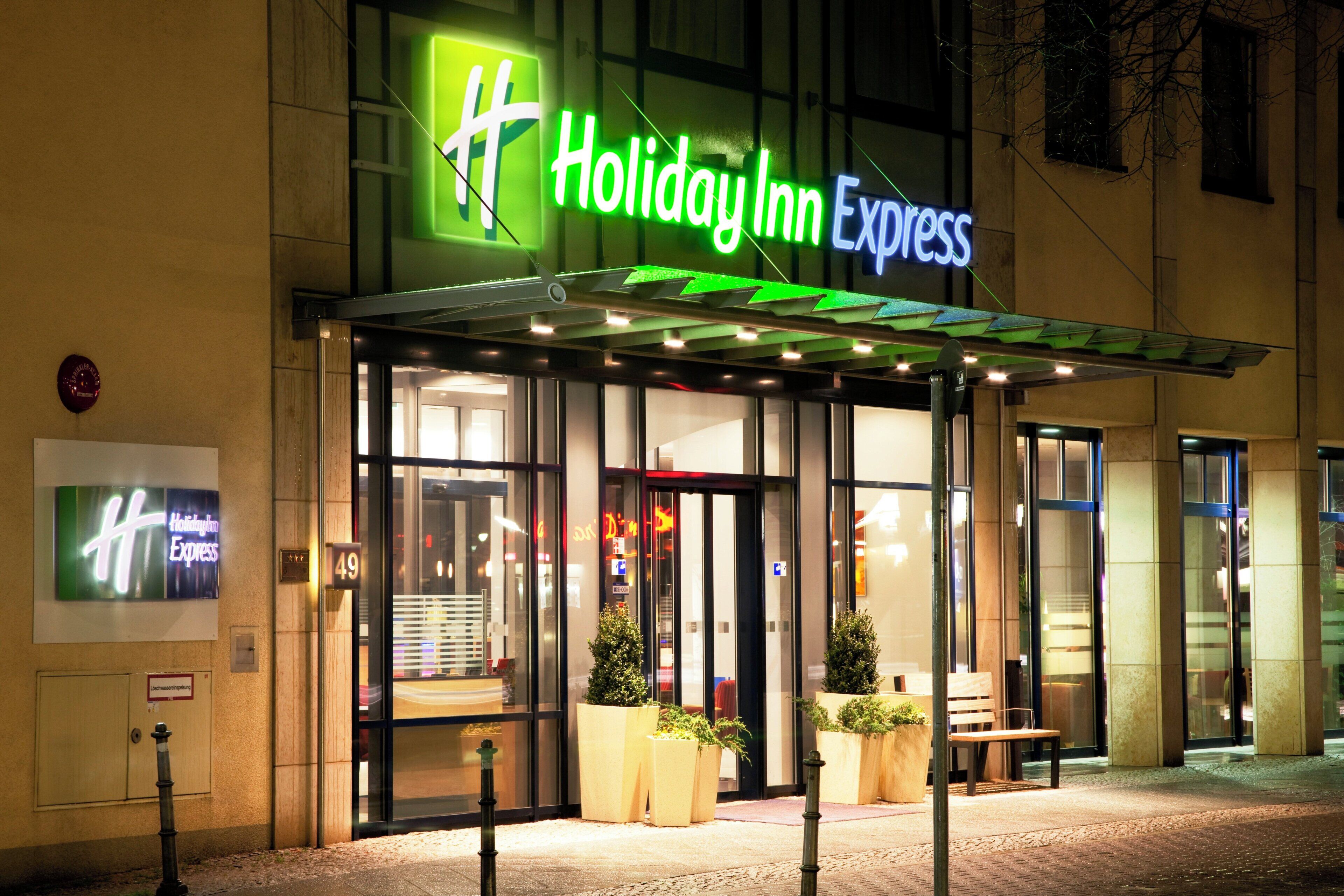 A Holiday Inn Express hotel entrance with a glass facade, outdoor seating, and a green neon sign.