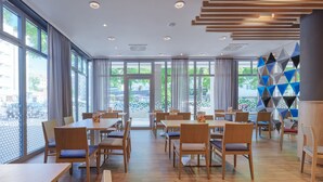 Un comedor moderno con mesas y sillas de madera, ventanas amplias con cortinas y una decoración geométrica en la pared.