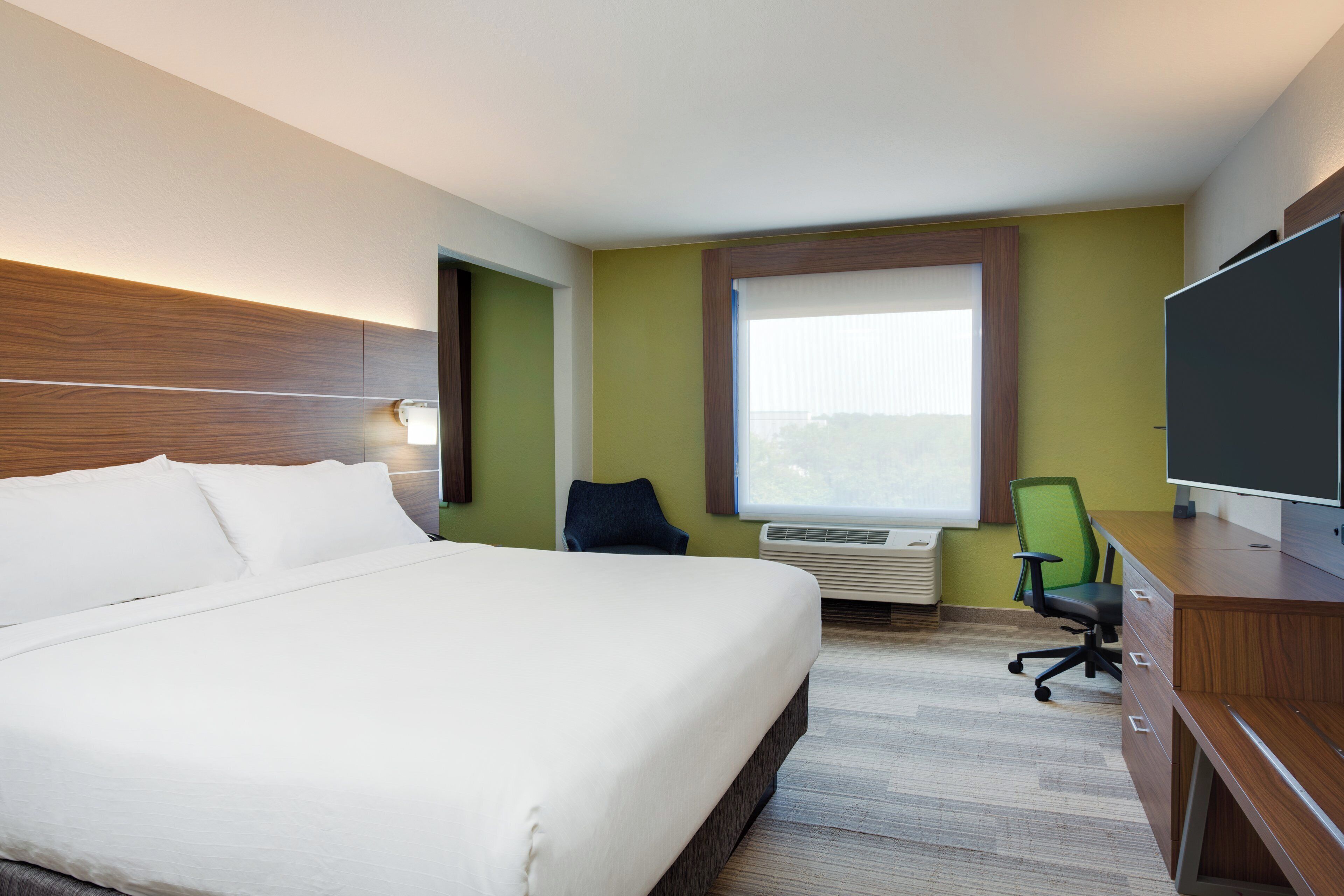 A hotel room with a large bed, a desk with a chair, an air conditioning unit, and a television.