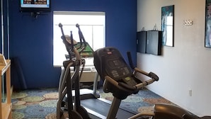 Un gimnasio con cintas de correr y elípticas, un televisor mostrando noticias económicas, y una alfombra estampada.
