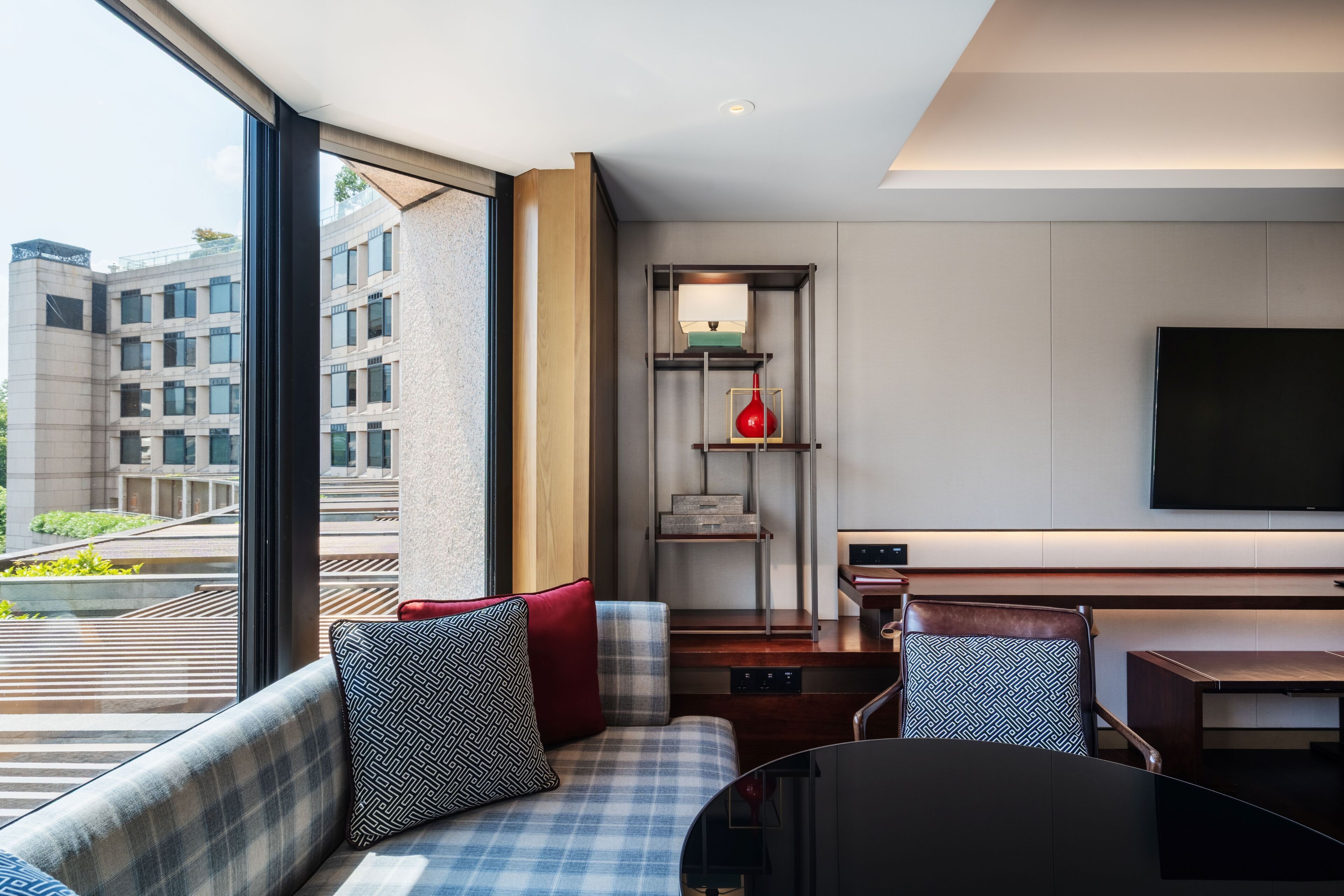 A modern hotel room with a large window, a sofa with patterned cushions, a dining table, and a flat-screen TV mounted on the wall.