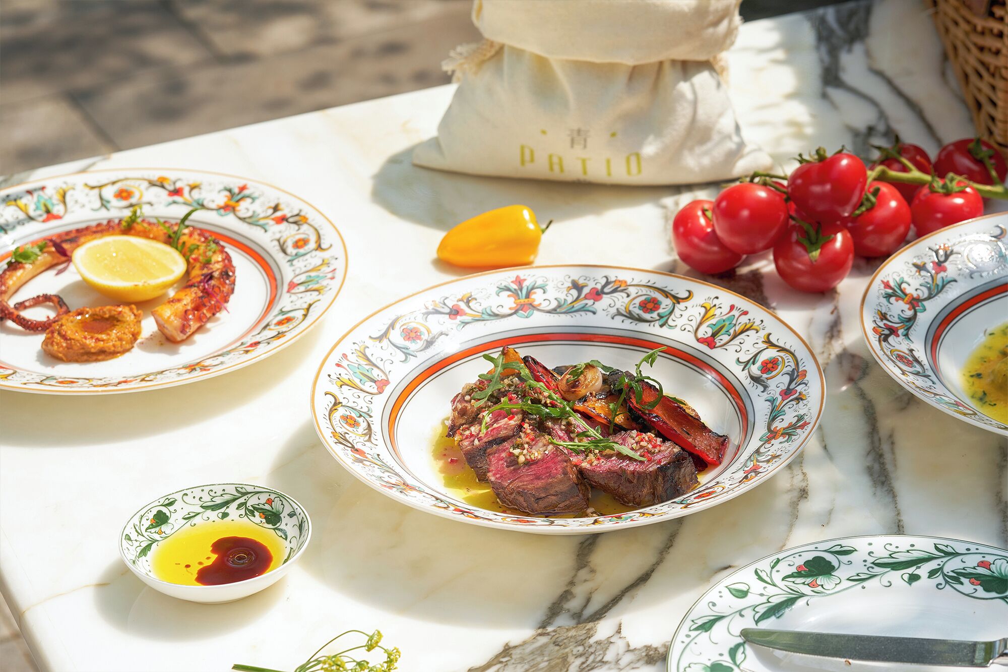 A table with plates of food, a bowl of sauce, and a basket of tomatoes.