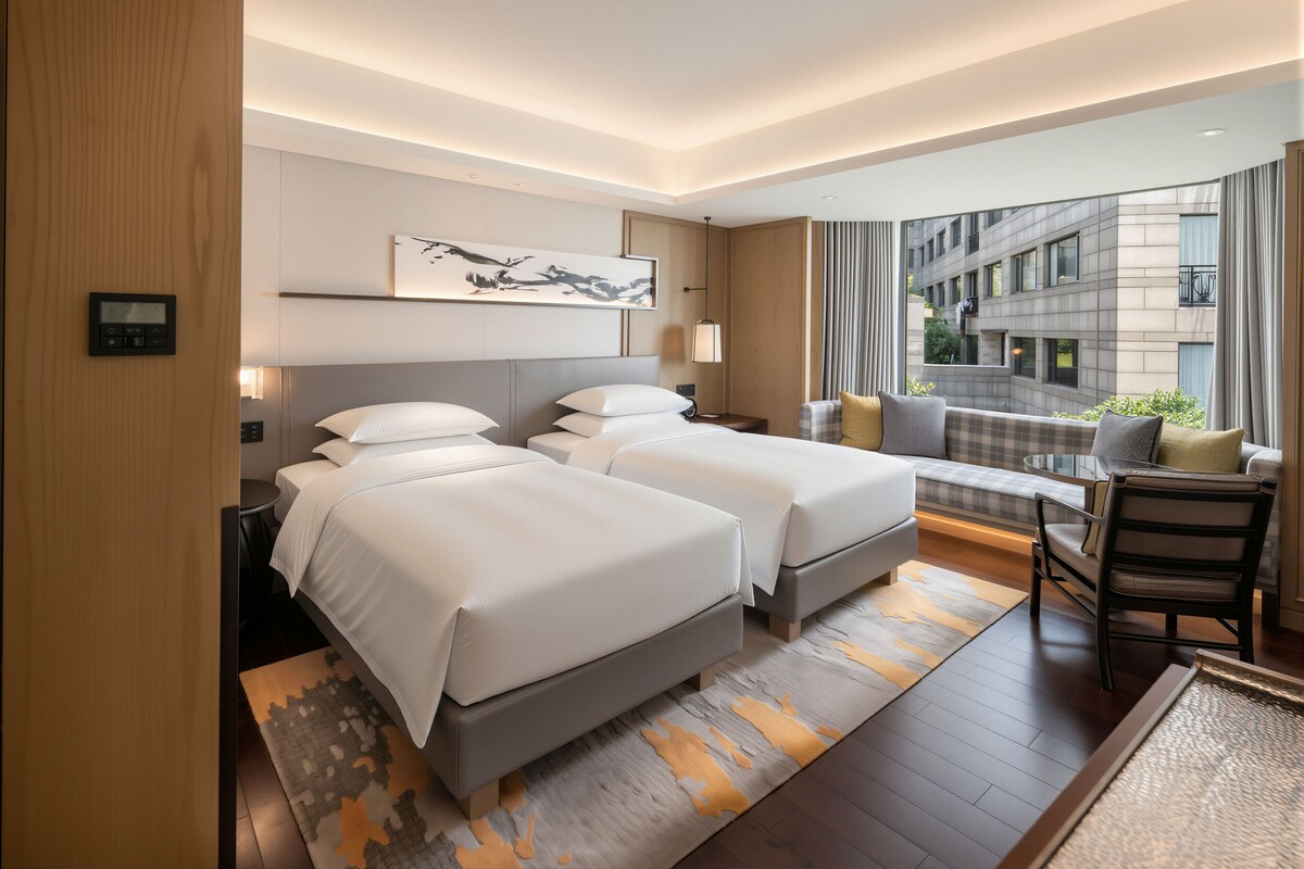 A hotel room with two beds, a desk, a chair, a large window with a view of buildings, and a painting on the wall.