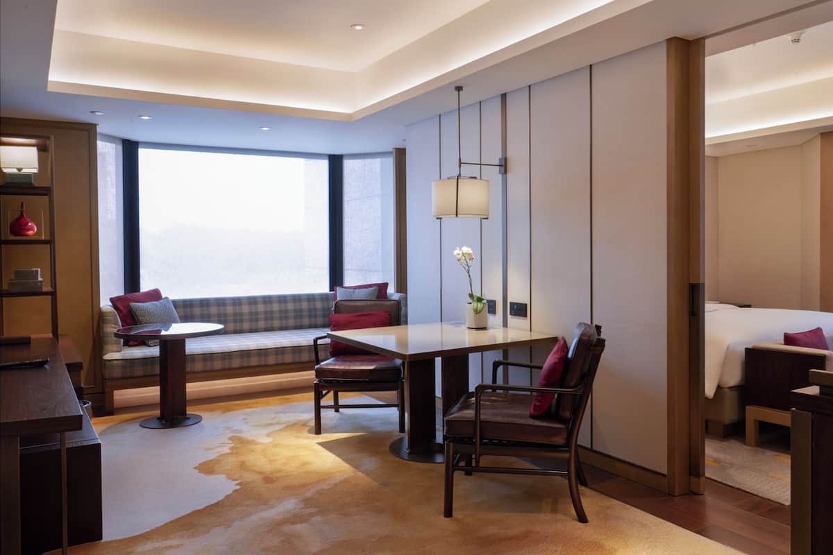 A modern hotel room with a large window, a sofa with cushions, a dining table, and a bed.