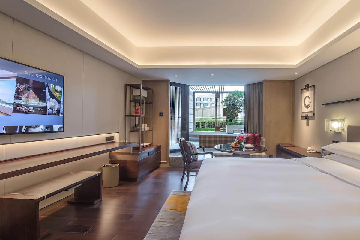 A modern hotel room with a large bed, a TV on the wall, a seating area with a table, and a view of the outdoors.