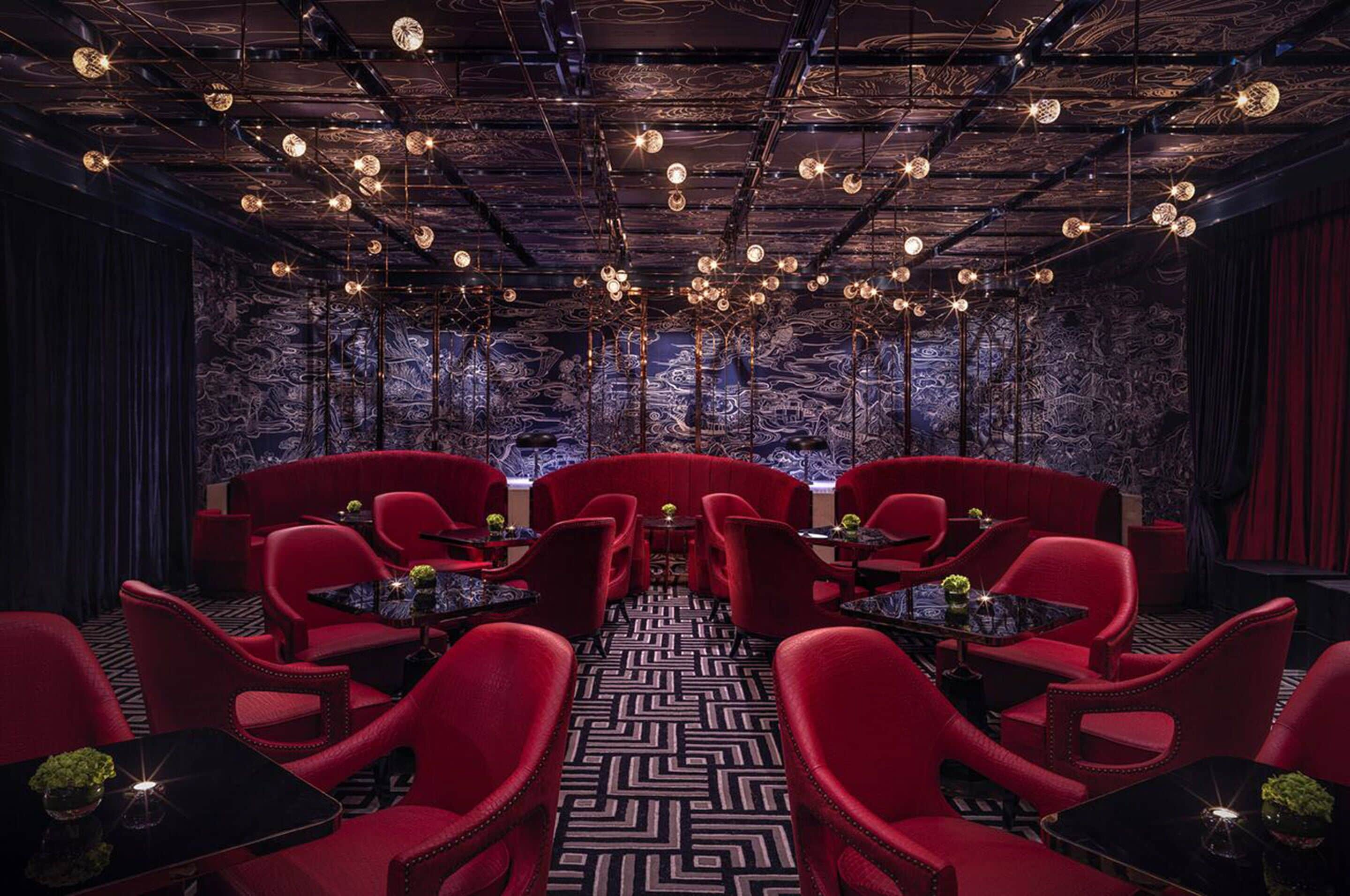 A room with red upholstered chairs and tables, a patterned floor, and a ceiling with exposed beams and hanging lights.