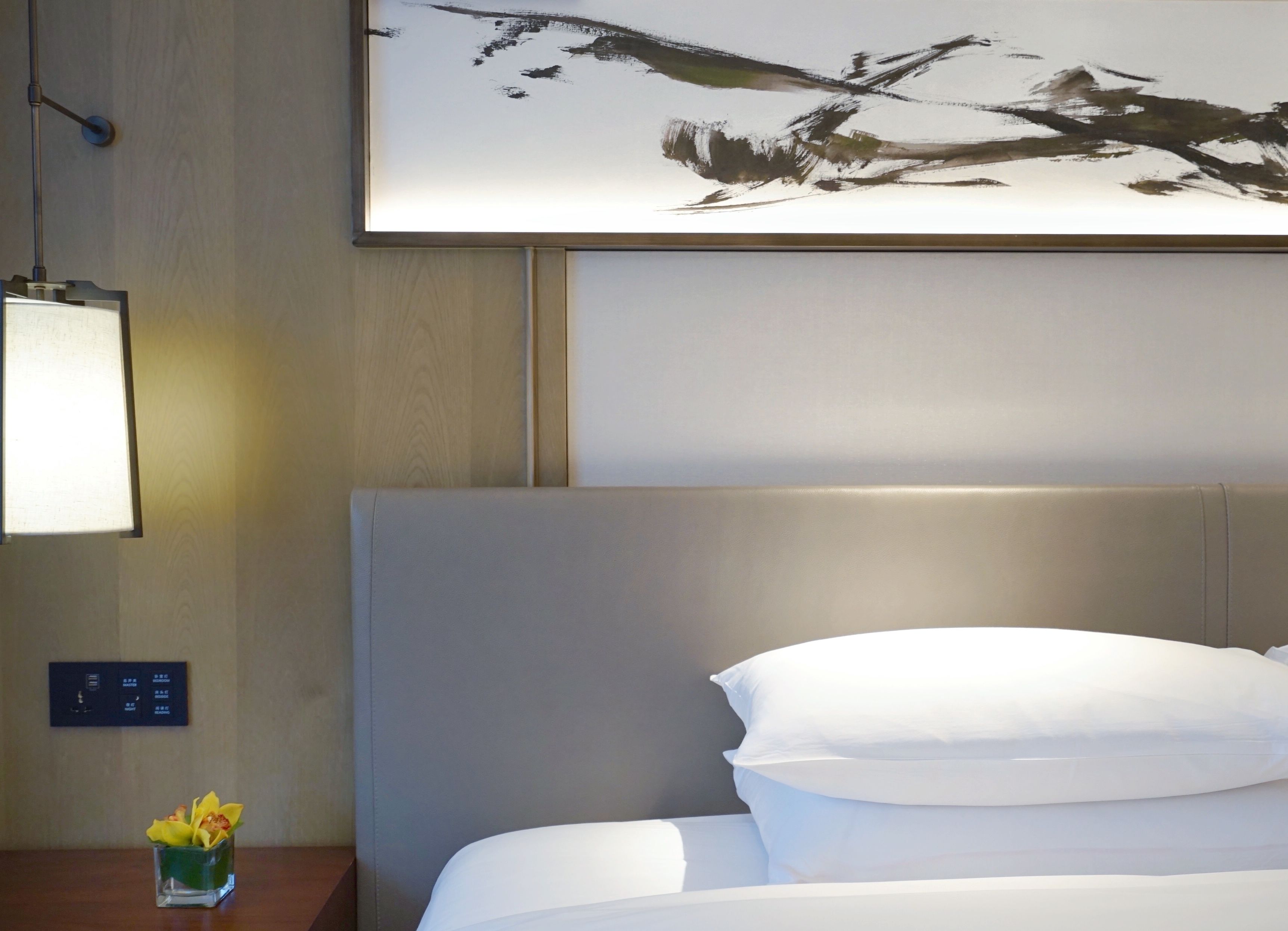 Habitación de hotel con cama, mesita de noche con lámpara, panel de control en la pared y un cuadro abstracto enmarcado sobre la cama.
