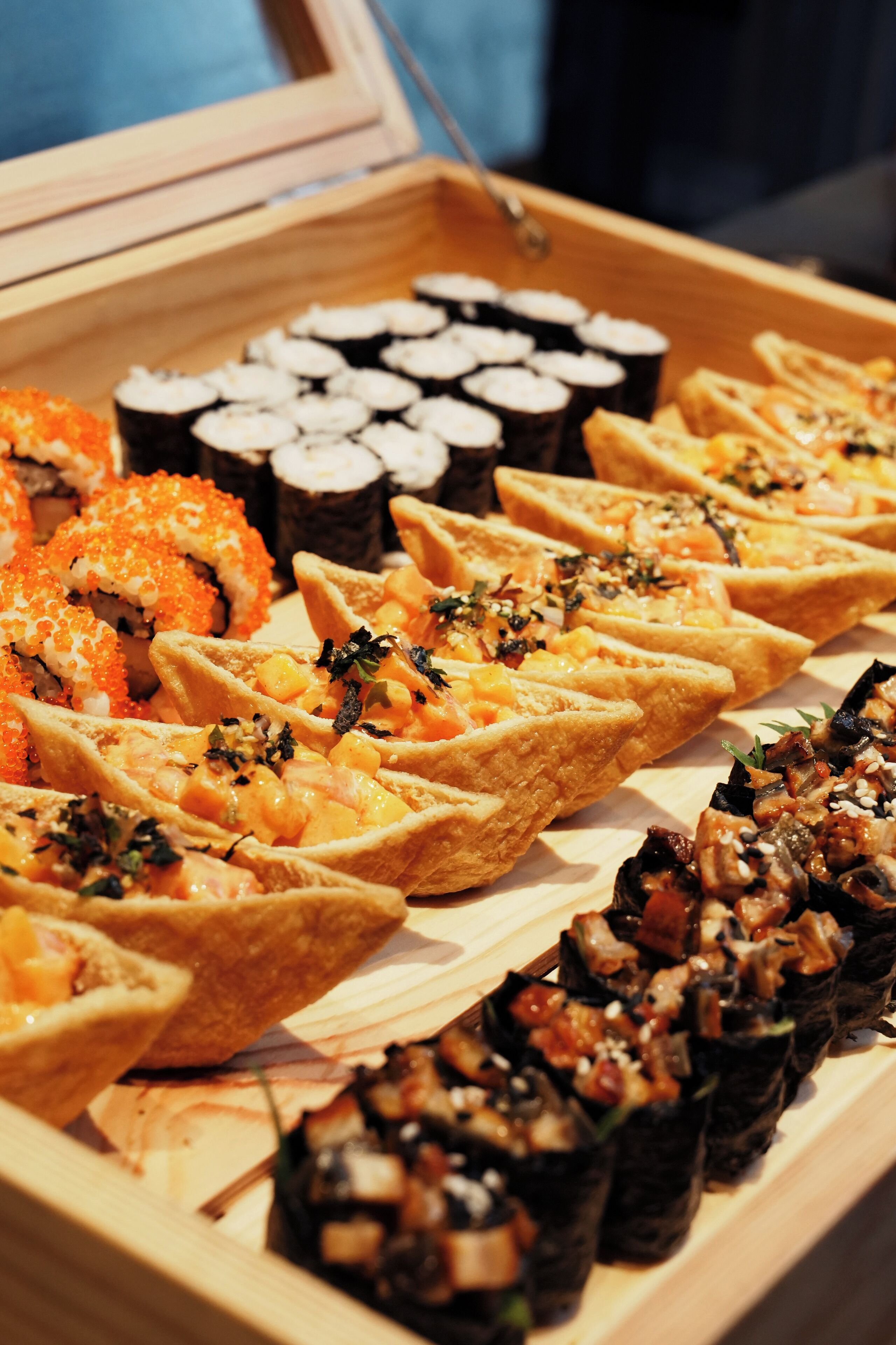A wooden tray with various sushi and appetizers, including rolls and topped with ingredients like fish roe and herbs.