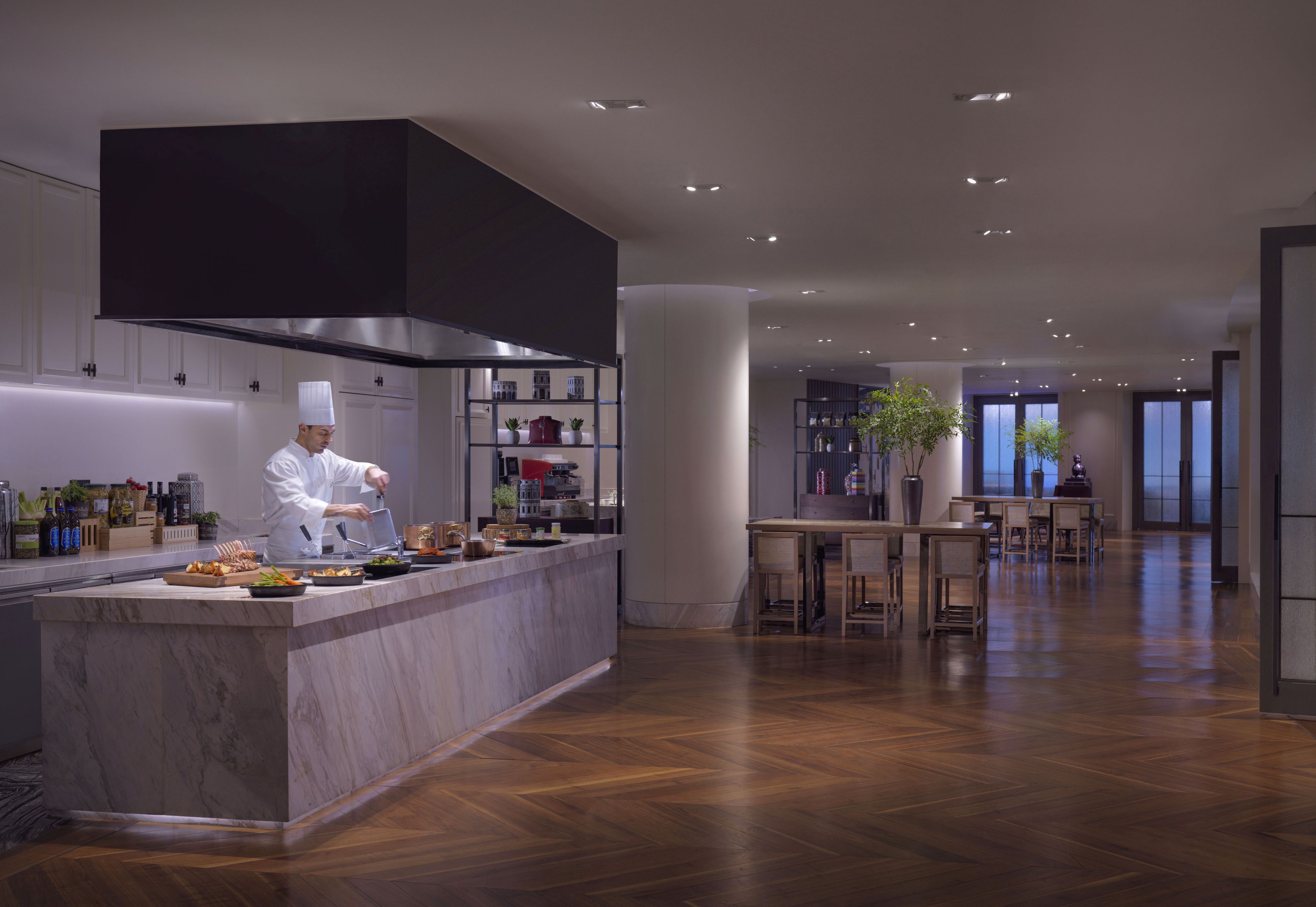 Cocina moderna con un chef preparando comida, encimera de mármol y zona de comedor con mesas y sillas de madera.