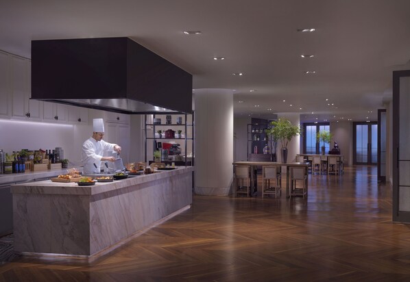 A modern kitchen with a chef preparing food, a marble countertop, and a dining area with wooden tables and chairs. - Grand Hyatt Hangzhou (Hangzhou)