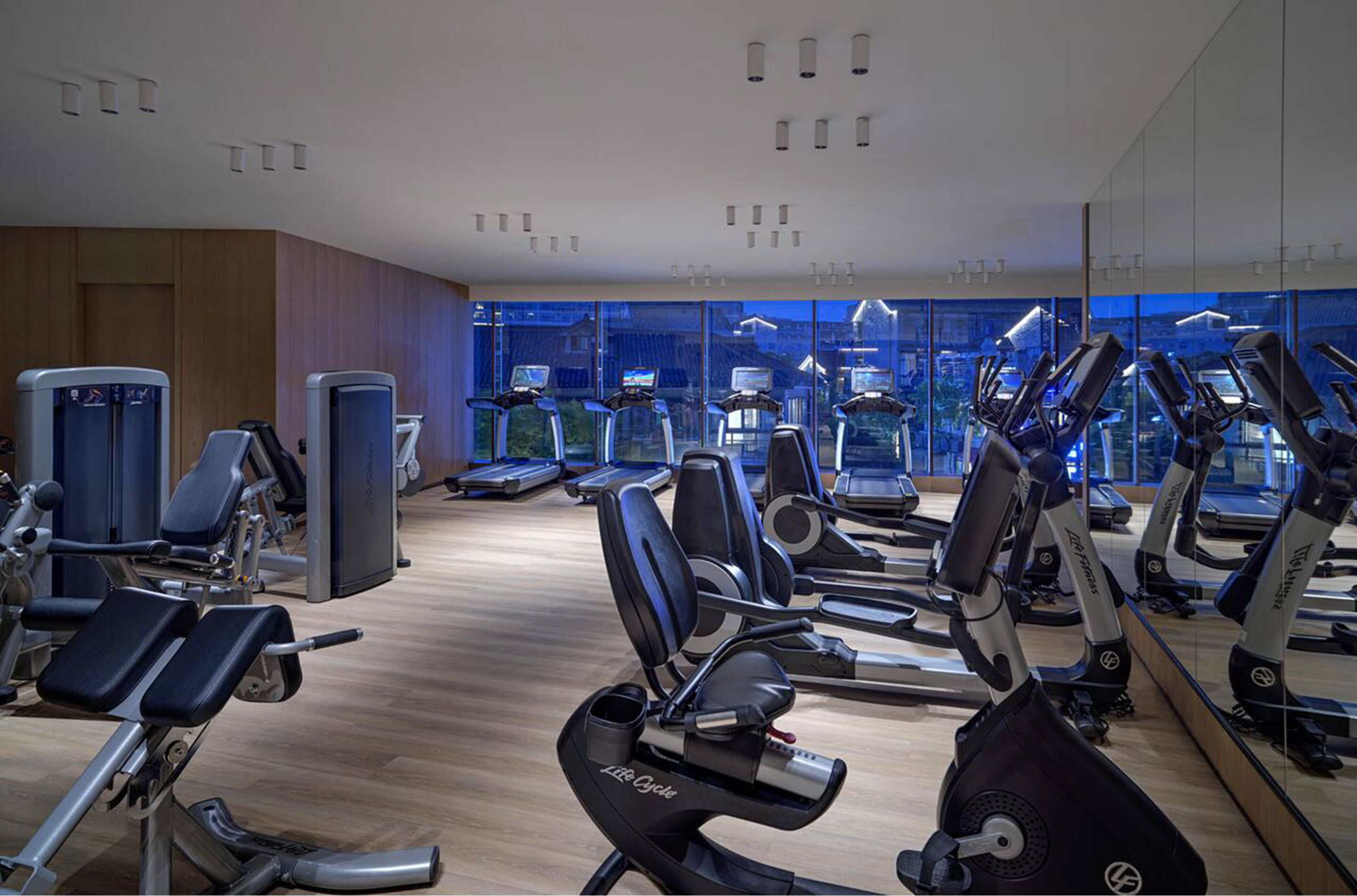 A gym with various exercise machines, a mirrored wall, and a view of the city through large windows.