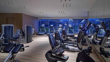 A gym with various exercise machines, a mirrored wall, and a view of the city through large windows.