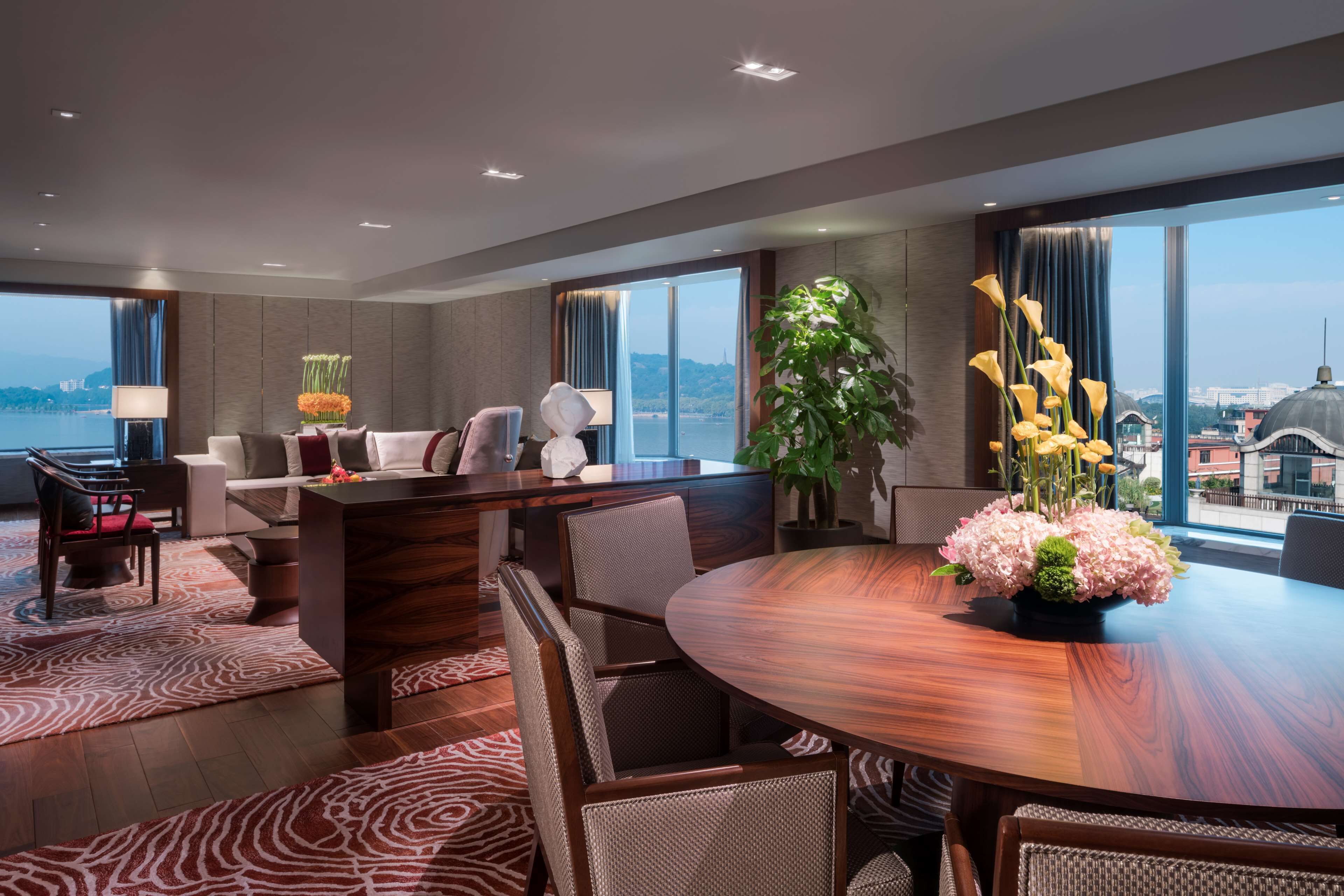 Une chambre d’hôtel moderne avec une grande table ronde, des sièges confortables, une grande fenêtre donnant sur la ville et une plante en pot.